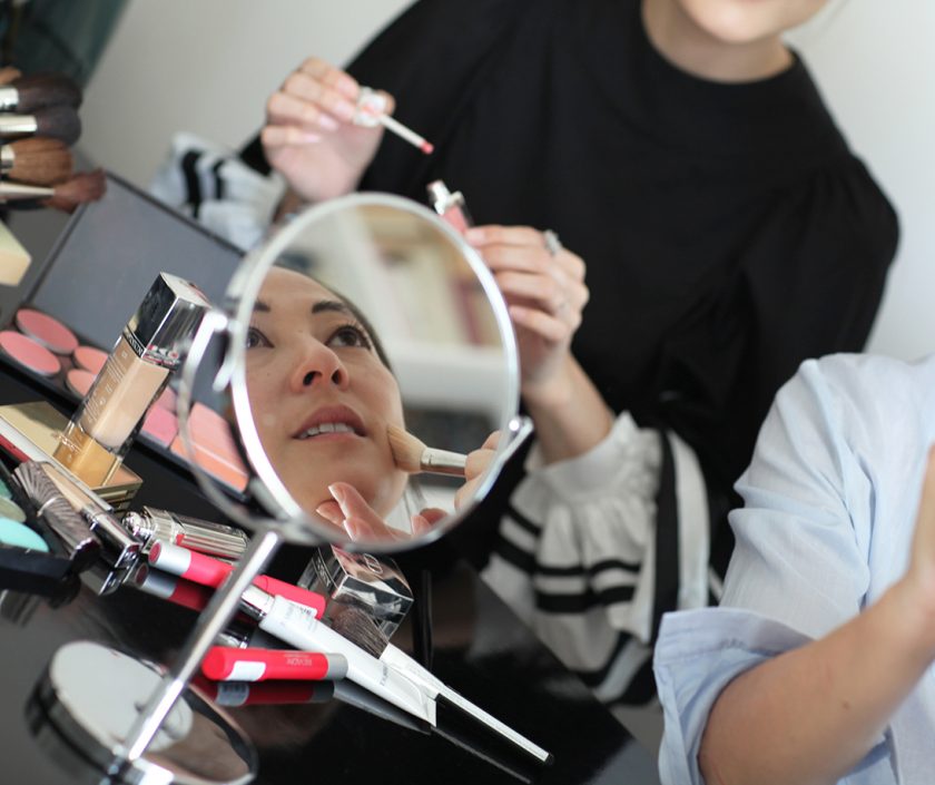 Un cours de maquillage entre copines. Une jeune femme asiatique, vu dans un miroir, est en train de se faire maquiller, par Louise Wittlich, maquilleuse pro et coach beauté
